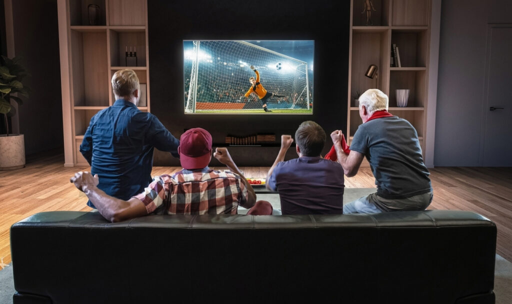 A képen egy négy tagú társaság látható, akik egy nappaliban, egy kanapén ülve néznek és szurkolnak a tv-ben szereplő futballmérkőzésnek.