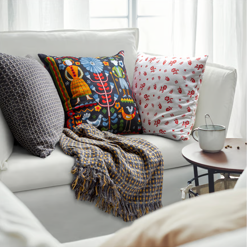 IKEA vintage stílusú díszpárna színes, népies mintával egy világos kanapén; mellette további párnák és mintás takaró, az előtérben kis kerek asztalka bögrével.